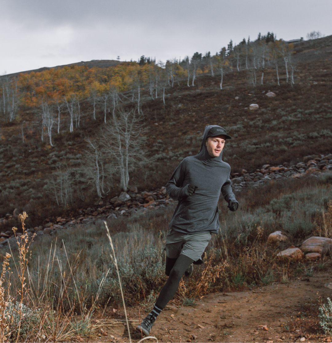 A man wearing a grey hoodie and cap runs on a dirt trail on a hillside.