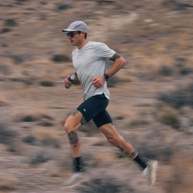 A man with tattoos runs on an outdoor trail, wearing a cap and holding a water bottle.