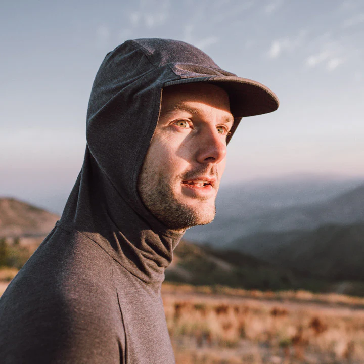 A man in a grey, brimmed hoodie looks to the side, outdoors in a hilly landscape.