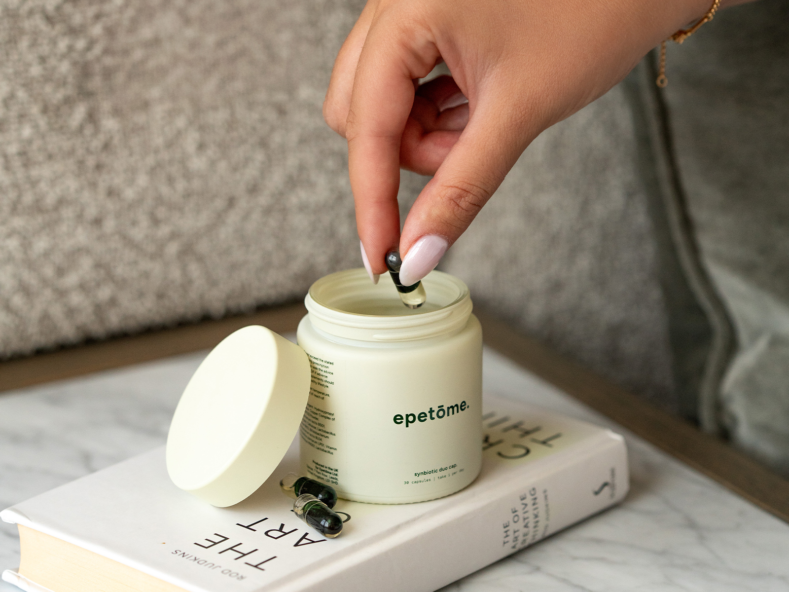 A hand with manicured nails takes a dark capsule from a cream-colored jar resting on a book.