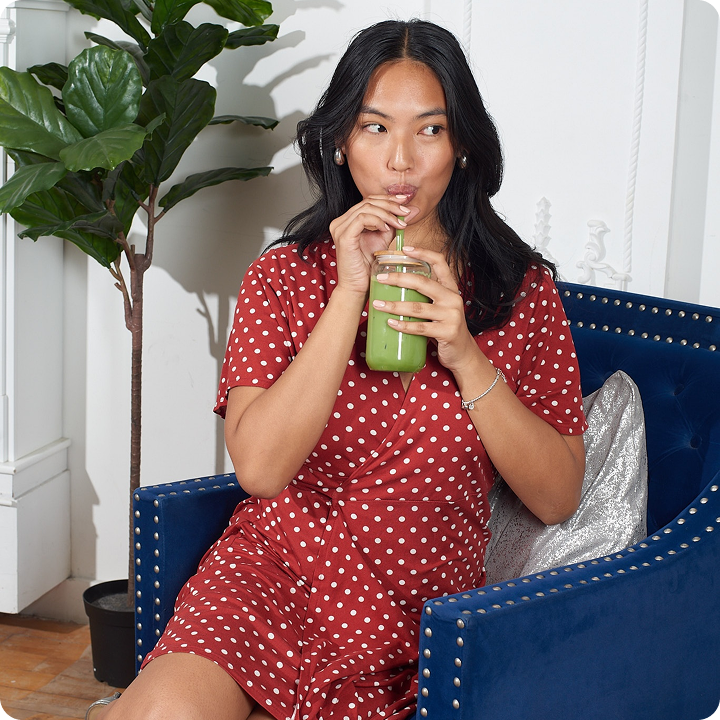 A woman in a red polka-dot dress sits in a blue chair, sipping a green drink through a straw.