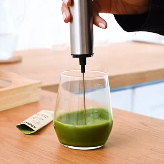 A person uses a handheld electric frother to mix a green beverage in a clear glass.