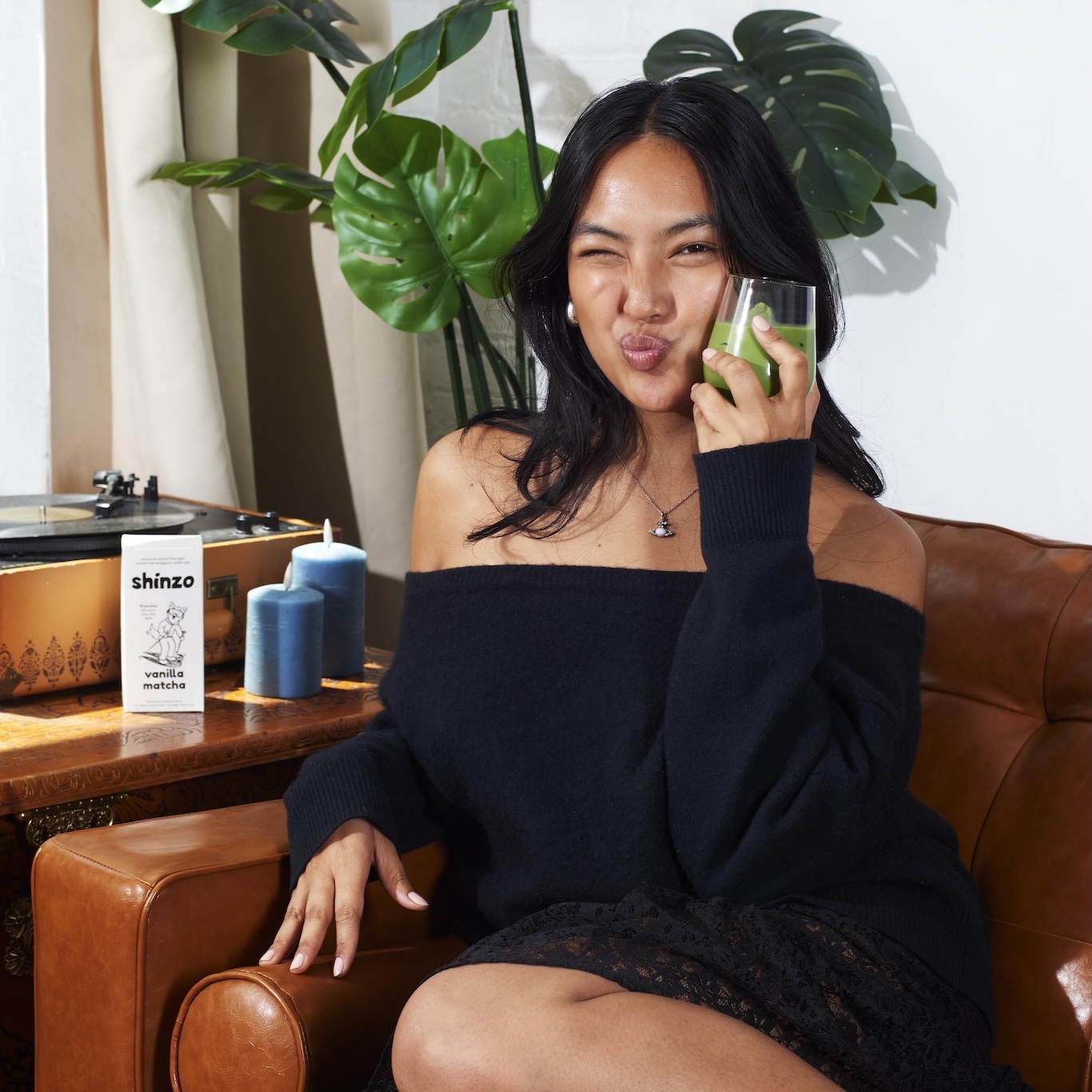 A woman in a black sweater winks while holding a glass of matcha, seated in a leather chair.