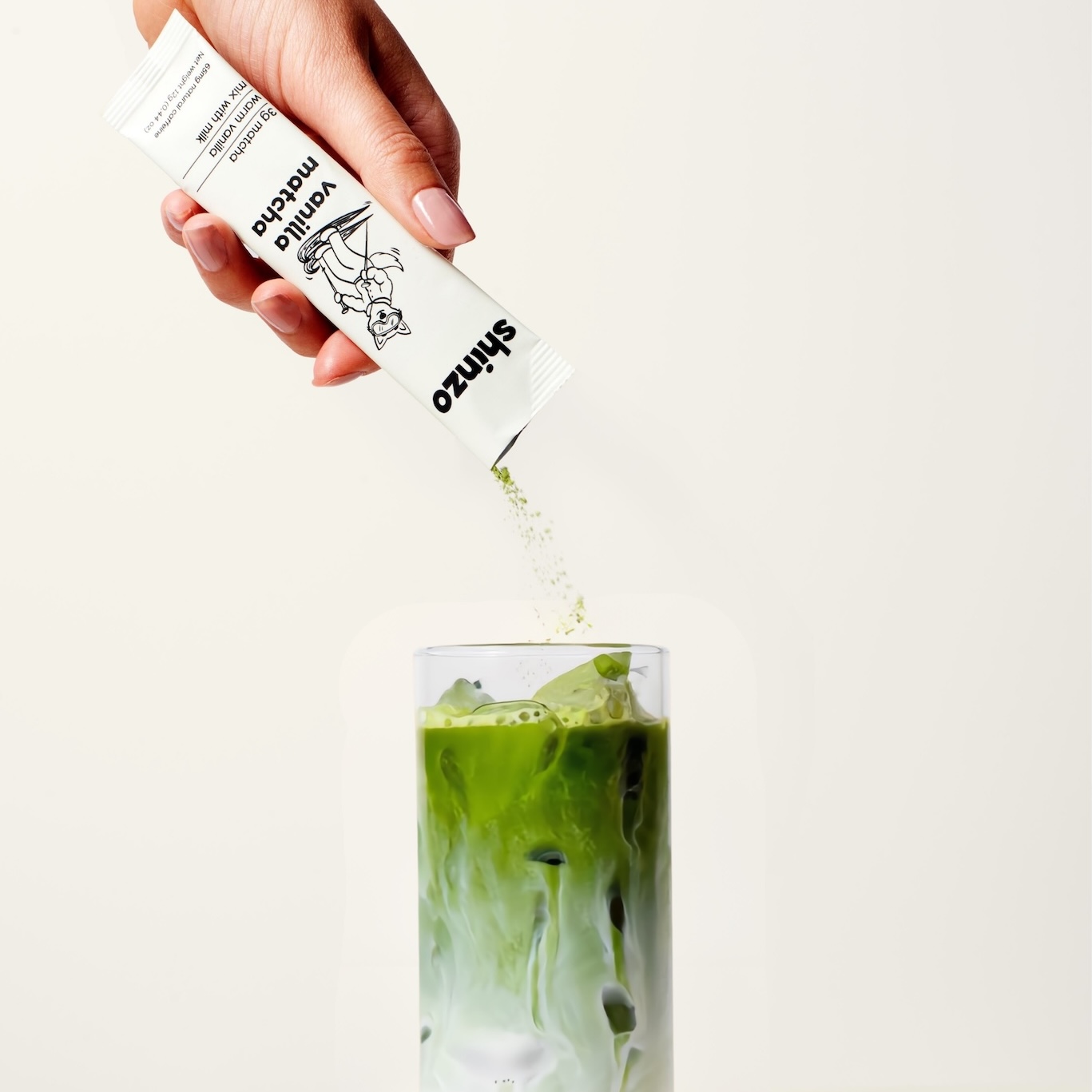 A hand pours vanilla matcha powder from a packet into a glass of iced matcha latte.
