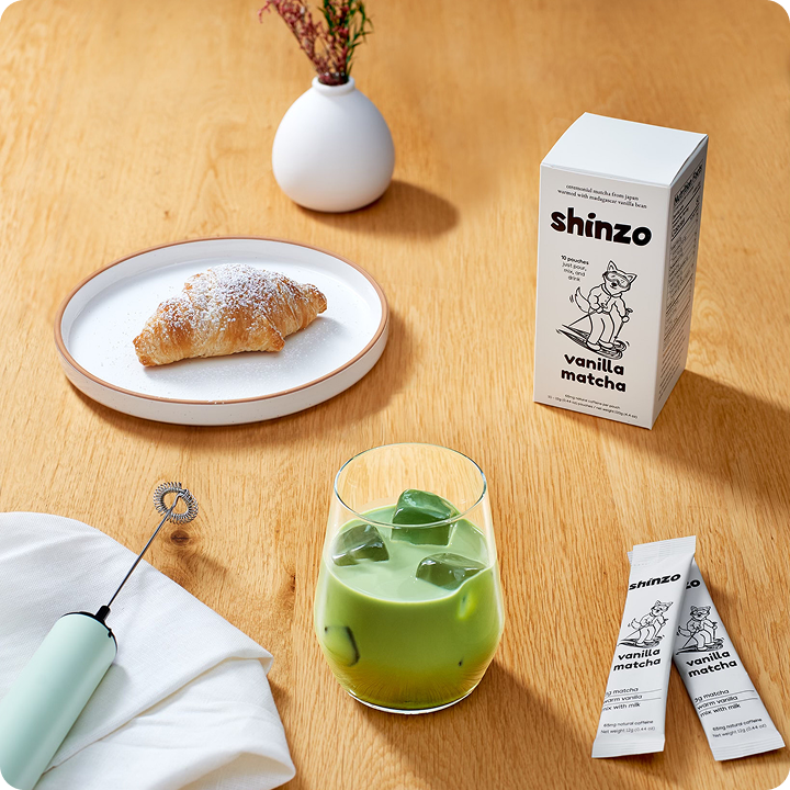 A glass of iced matcha, a croissant, and Shinzo vanilla matcha packaging on a wooden table.