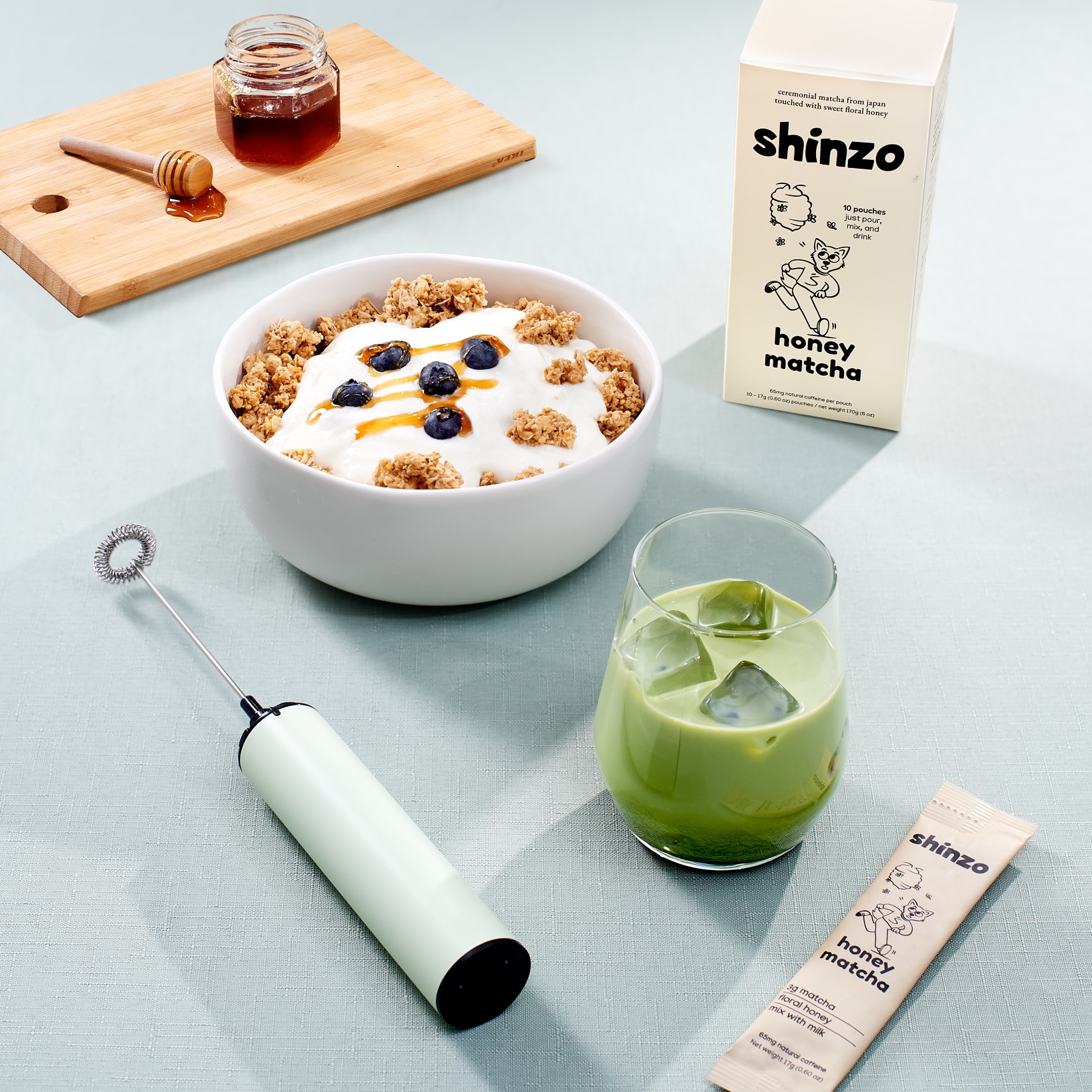 A bowl of granola and yogurt, a glass of iced matcha, a frother, and honey matcha mix.