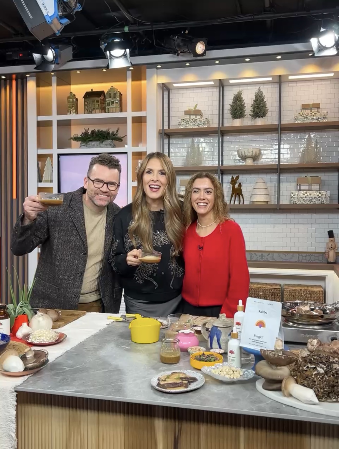 A man and two women smile and hold up small cups on a TV cooking show set.