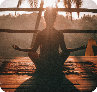 A silhouette of a person in a meditation pose on a wooden deck at sunrise or sunset.