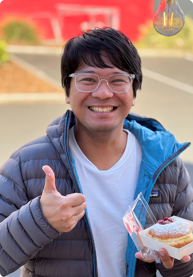 A smiling man in a puffer jacket holds a jelly donut and gives a thumbs up.