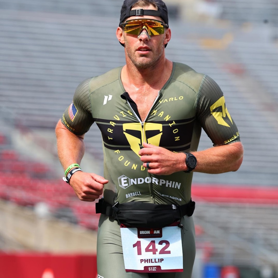 A male triathlete wearing sunglasses and a green race suit runs during an Ironman event in a stadium.
