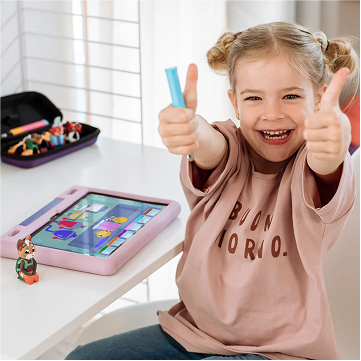 Smiling child giving thumbs up next to a tablet on a table with toys.