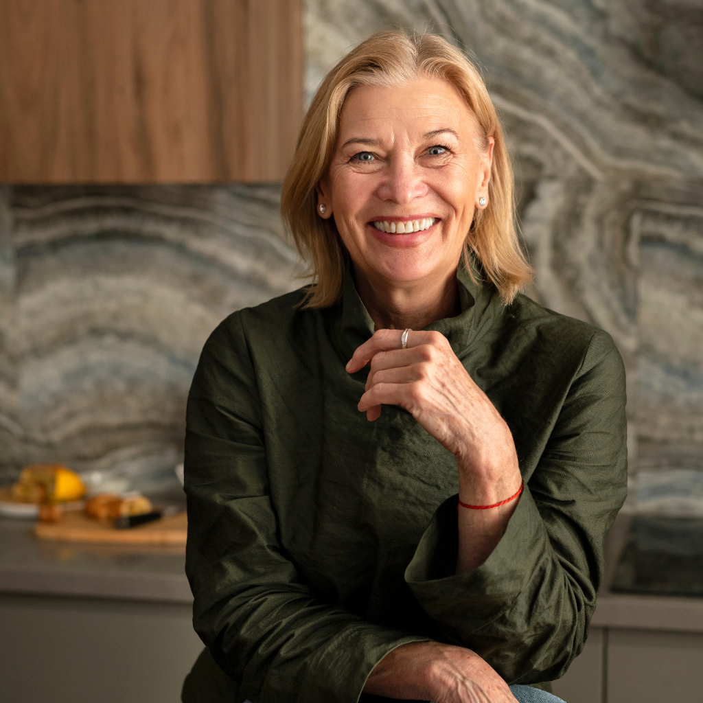 A smiling, older blonde woman in a green shirt poses for a portrait in a modern kitchen.