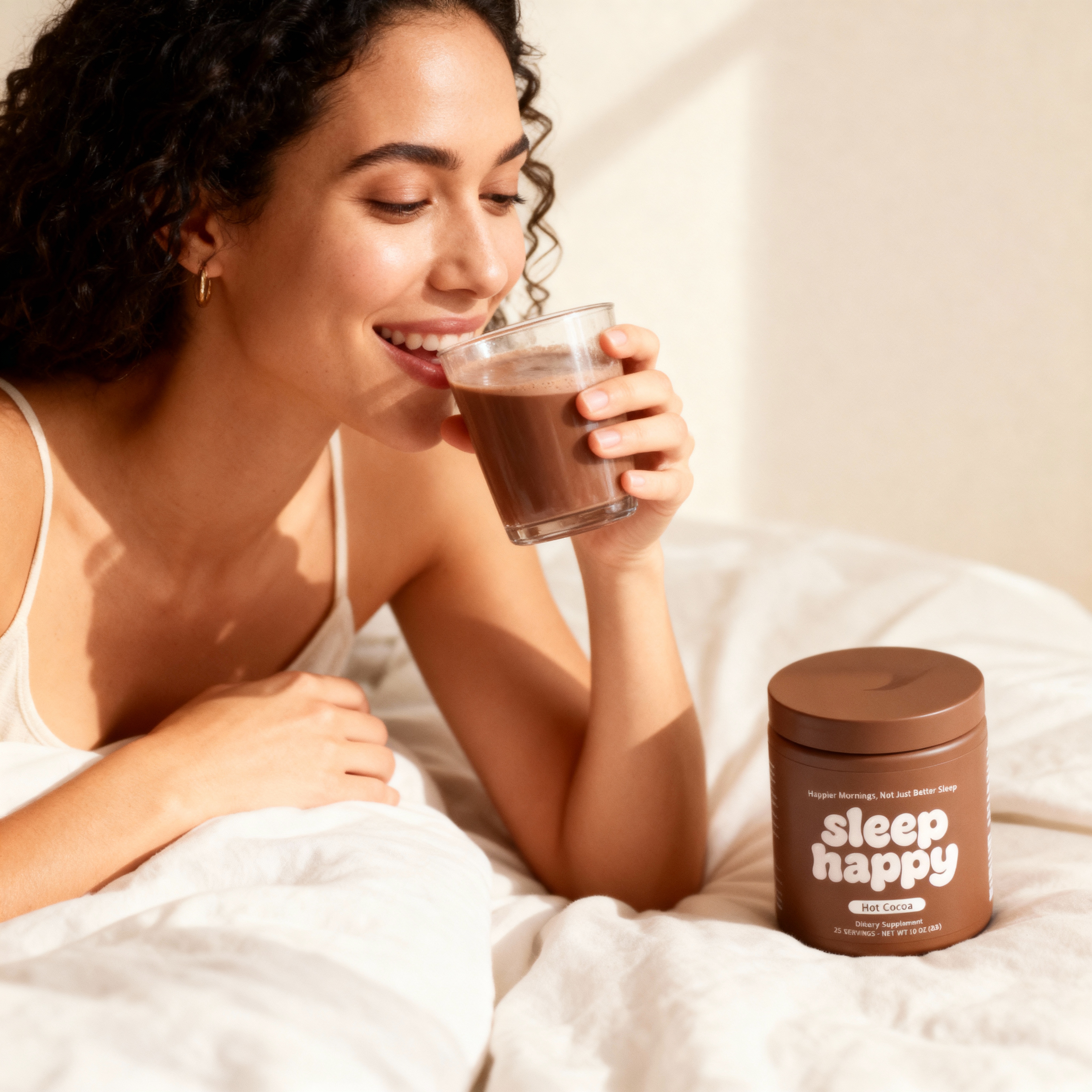 Person in bed drinking cocoa from a glass, beside a cocoa container.