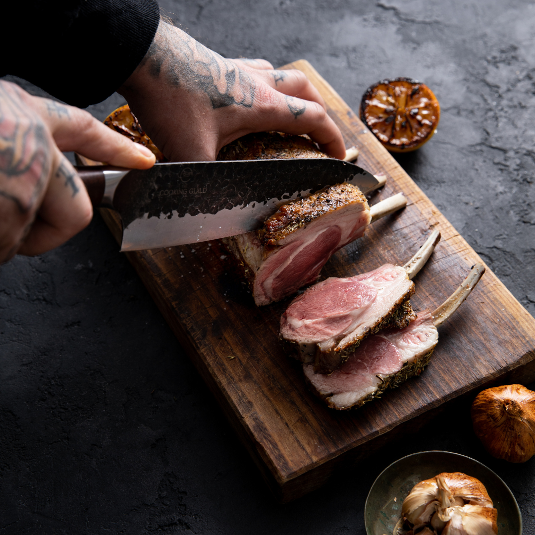 Person slicing cooked rack of lamb on a wooden cutting board.