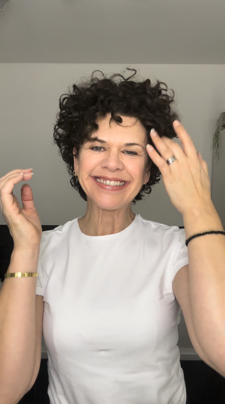 Smiling person with curly hair wearing a white shirt, raising hands near face.