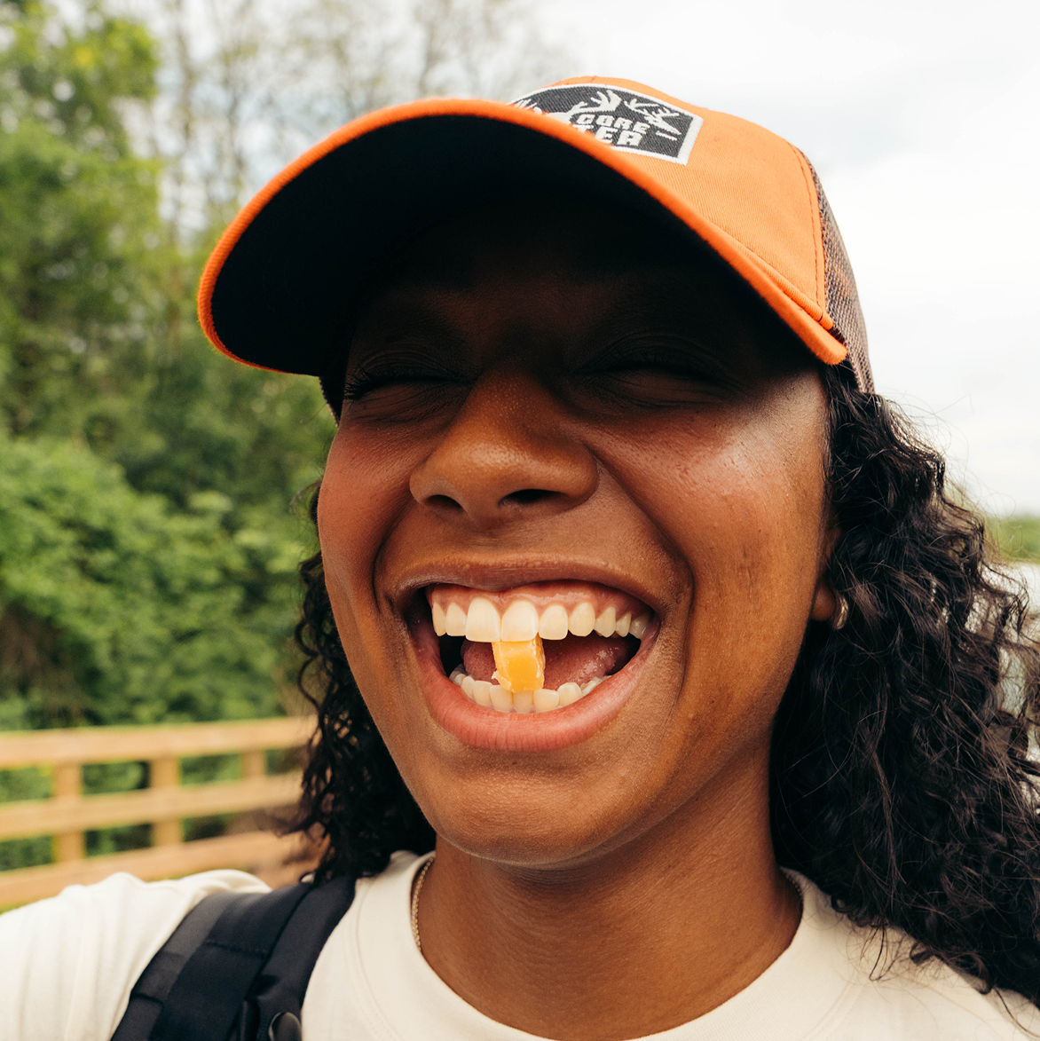 Person smiling with a piece of food between teeth, wearing an orange cap outdoors.