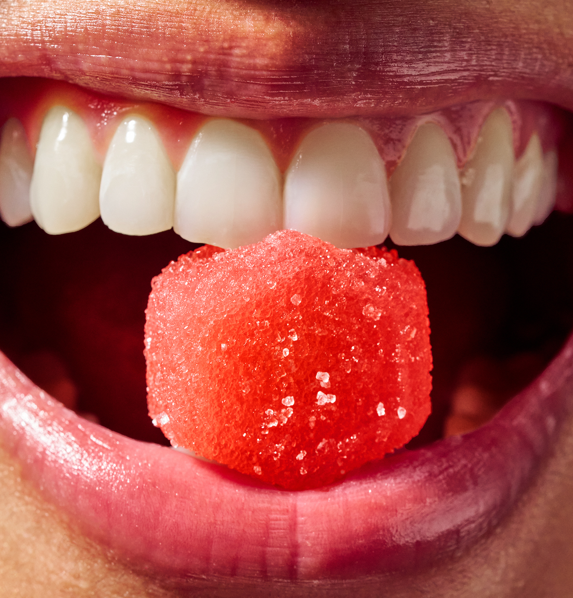 Close-up of a mouth holding a red sugar cube between teeth.