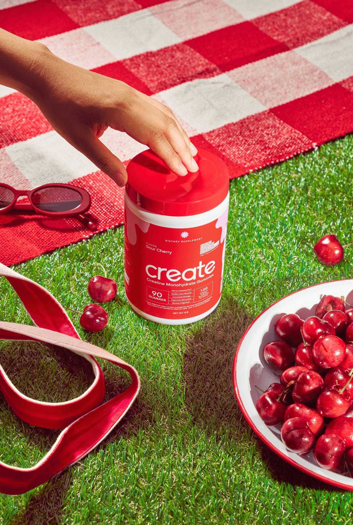 Hand reaching for a red container on grass, with cherries and sunglasses nearby.