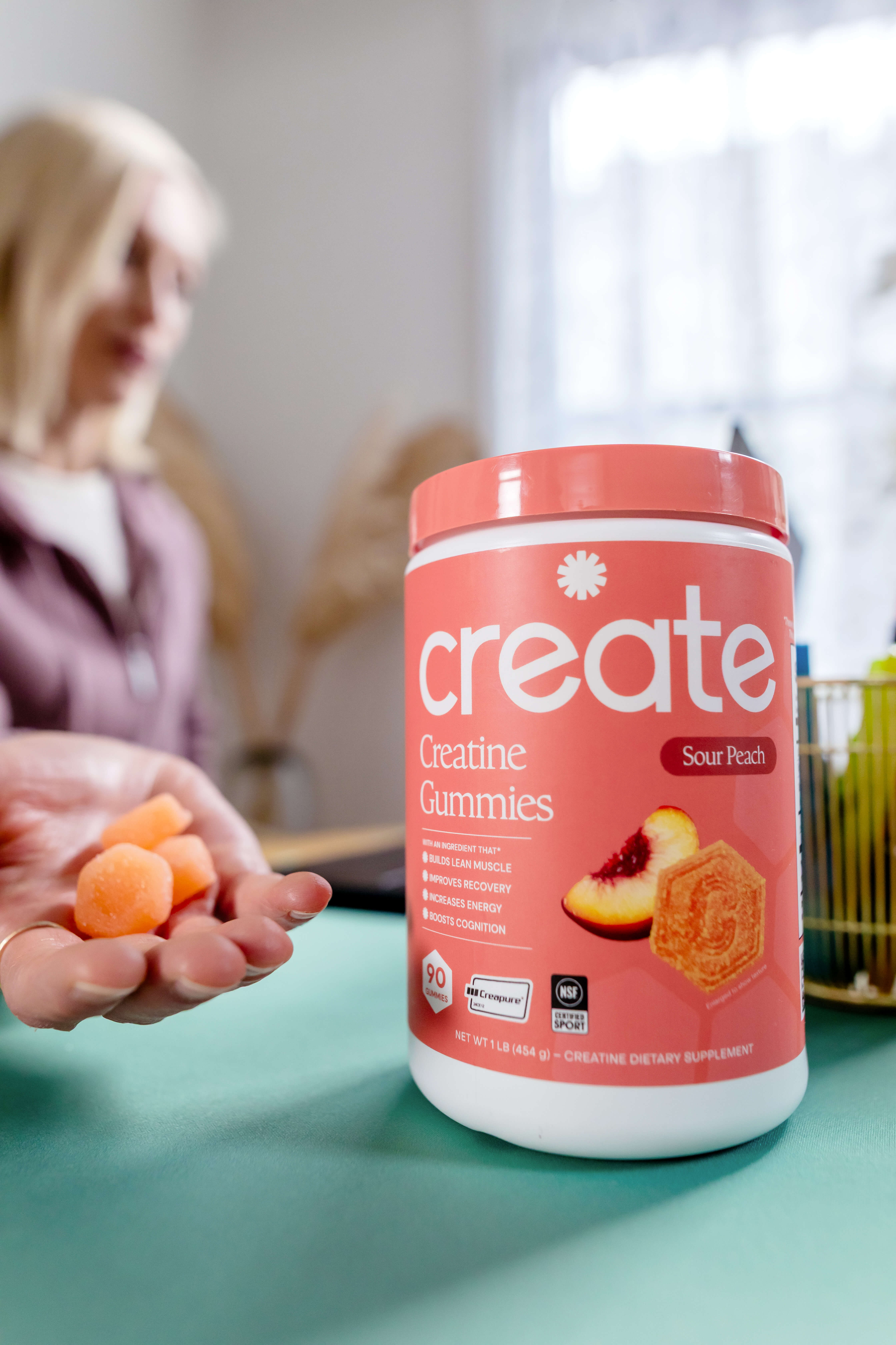 A container of Create Creatine Gummies sits on a table, while a person holds a few gummies.
