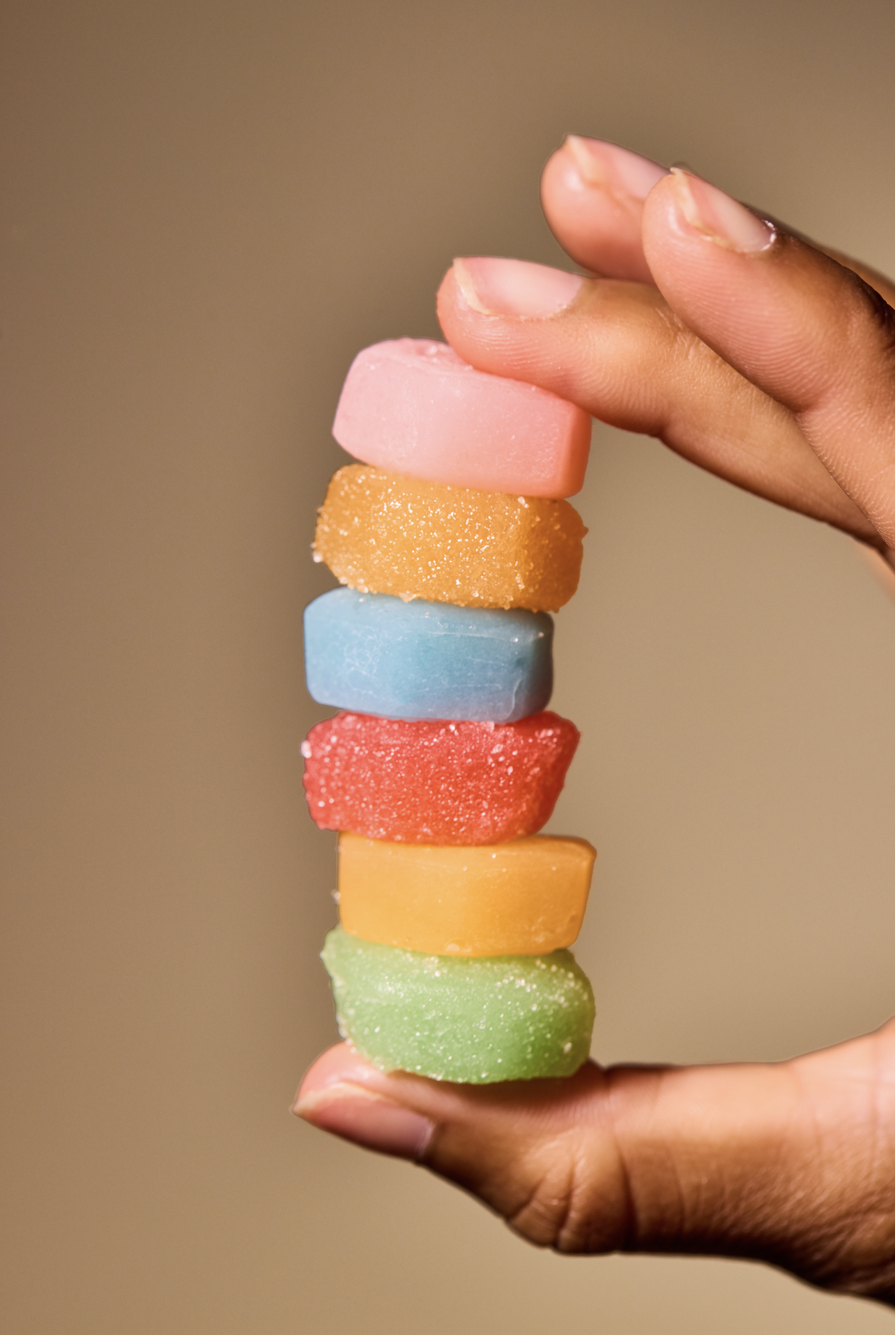 A person's hand holds a vertical stack of six colorful, sugar-coated, square-shaped candies against a plain background.