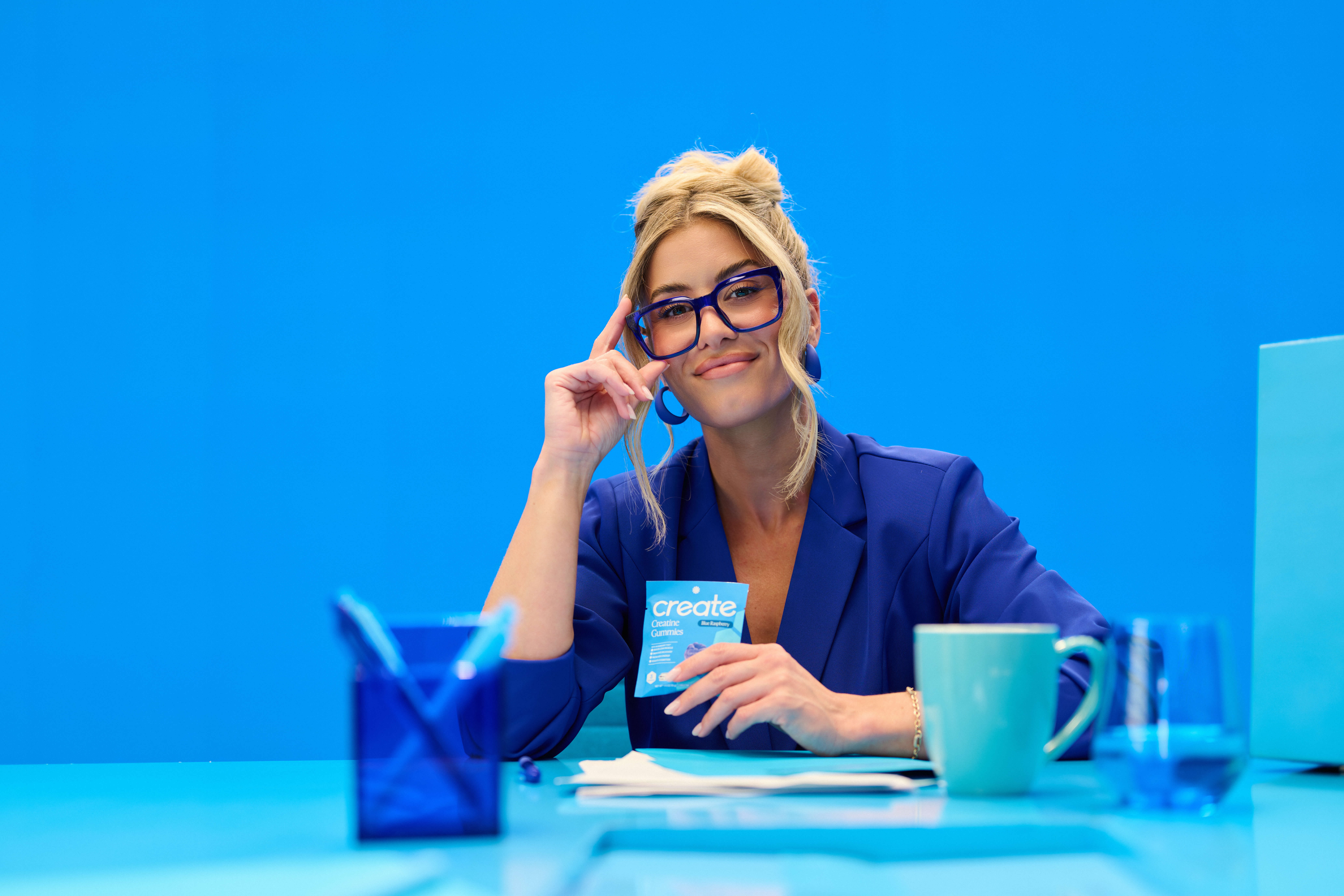 A woman in a blue blazer and glasses sits at a desk holding a pouch of creatine gummies.