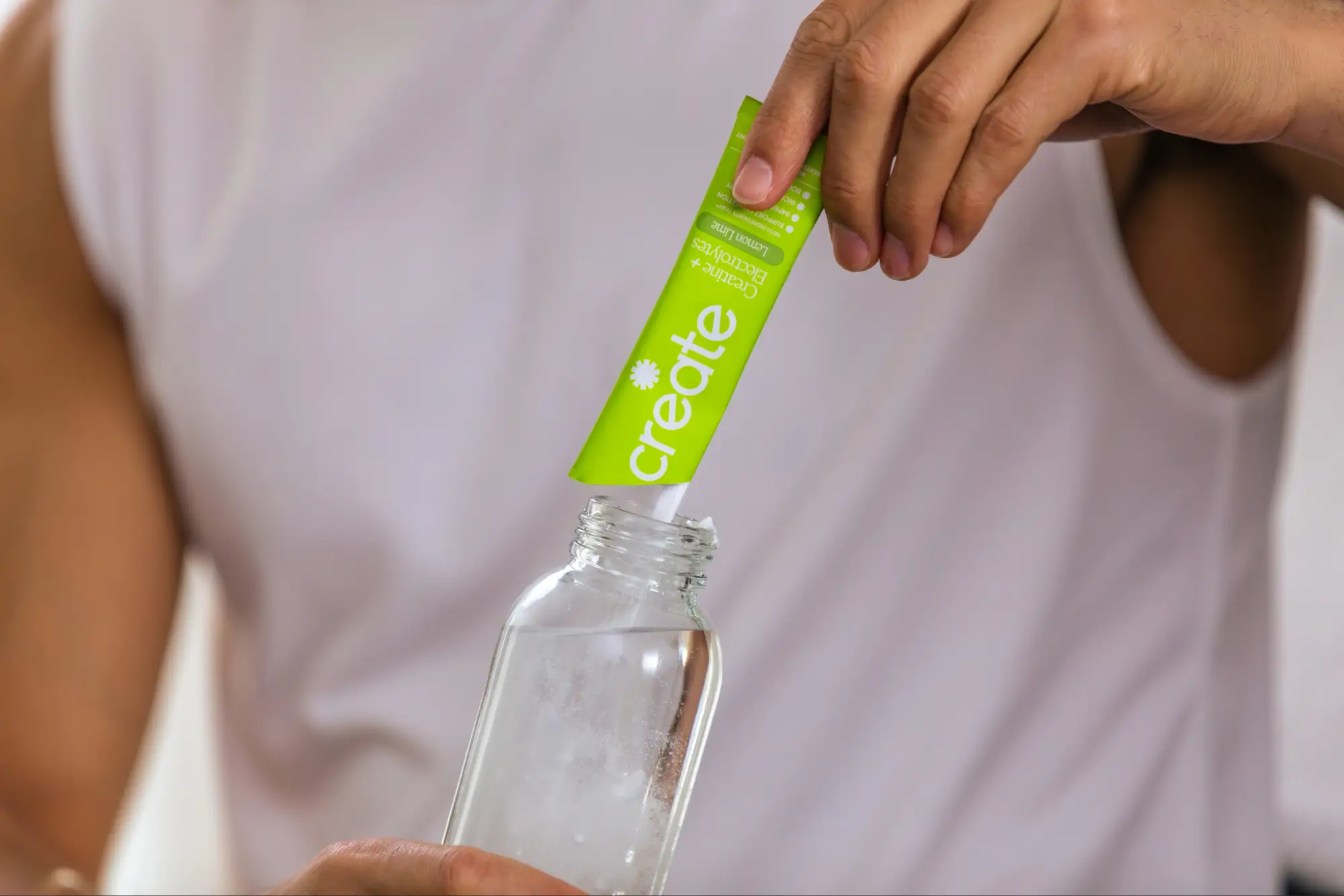 A person pours a green packet of Create Creatine + Electrolytes powder into a clear glass bottle.