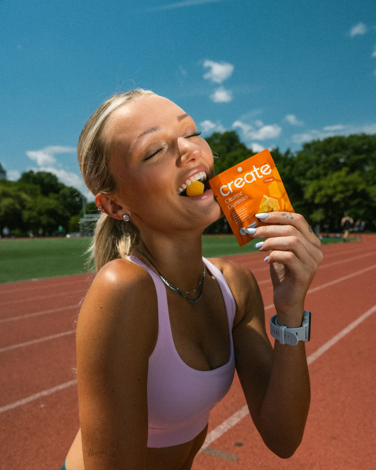 A woman on a running track biting into a Create gummy from an Orange pack