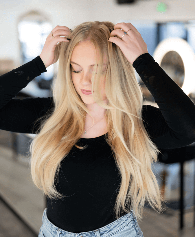 Blonde woman in a black top running her fingers through long, voluminous Bello Haven hair extensions in a salon setting
