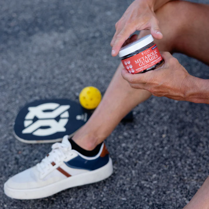 Person holding a jar of metabolic gummies, with paddle and ball on the ground.