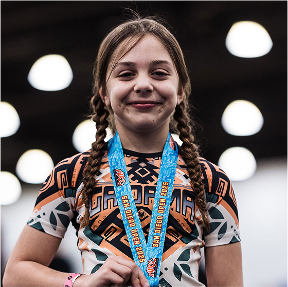Young girl with braided hair wearing a medal and patterned shirt, smiling indoors.