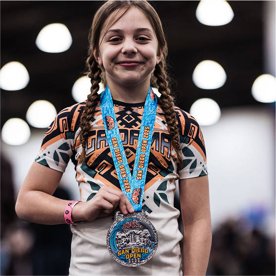 Young girl with braided hair wearing a medal and patterned shirt, smiling indoors.