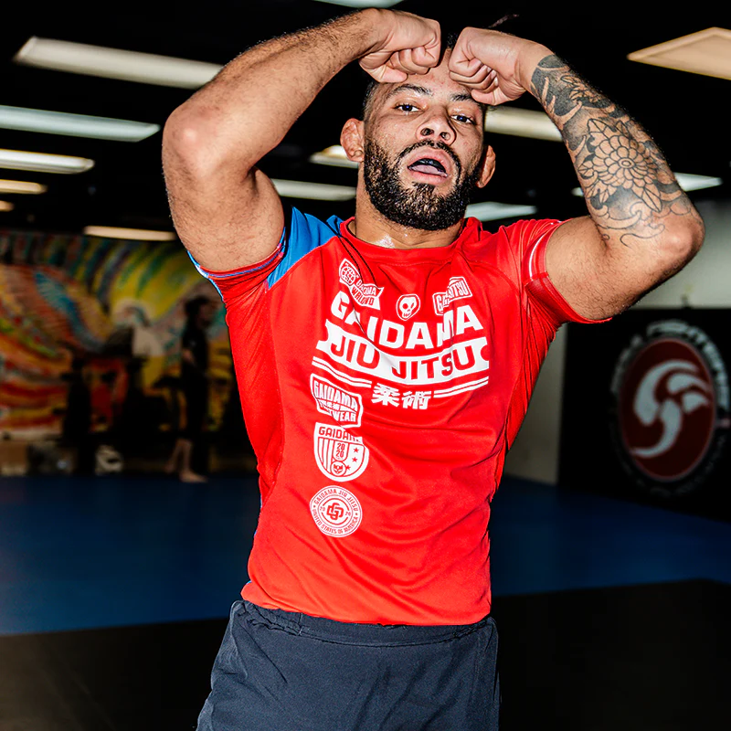 Man in red jiu-jitsu shirt with tattoos, posing in a gym.