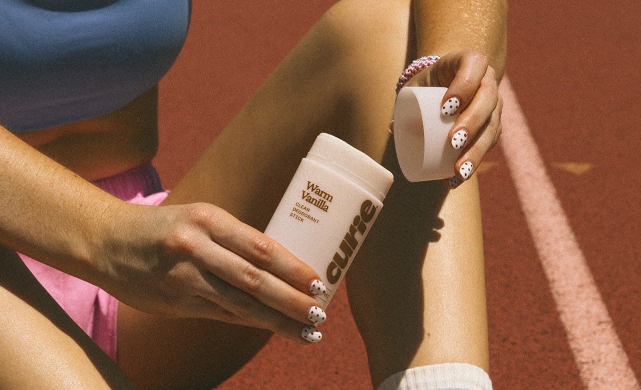 A person holds a stick of Curie Warm Vanilla deodorant while sitting on a red sports court.