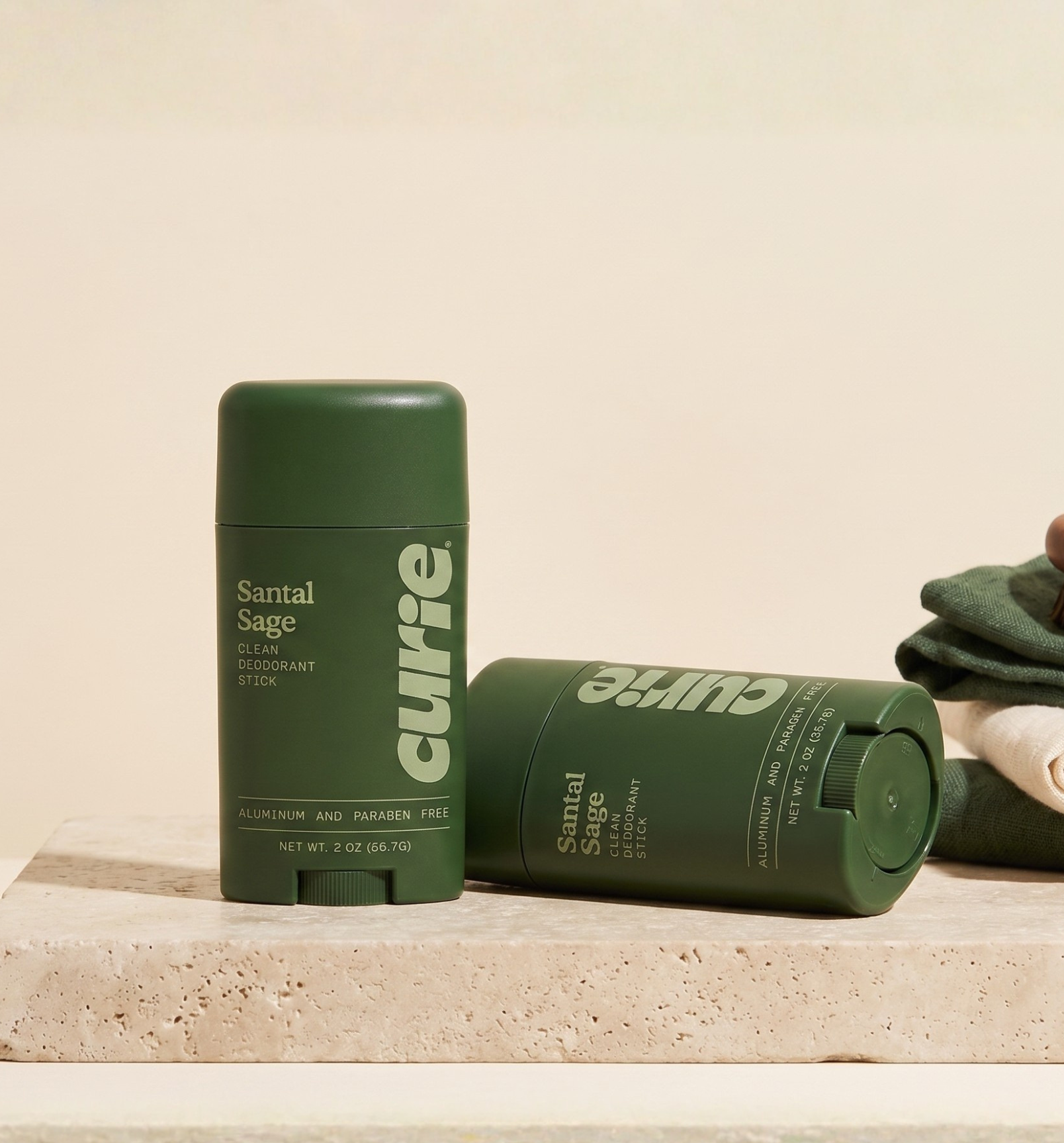 Two green sticks of Curie Santal Sage deodorant, one upright and one on its side on a stone slab.