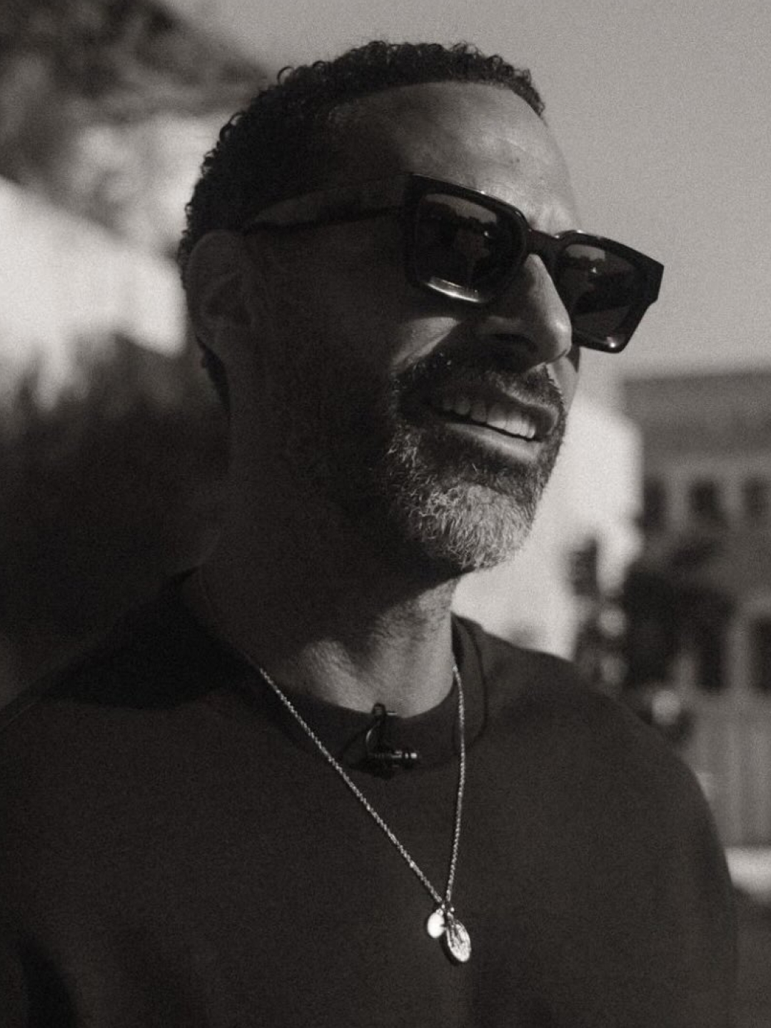 A black and white photograph of a smiling man with a beard, wearing sunglasses and a necklace.