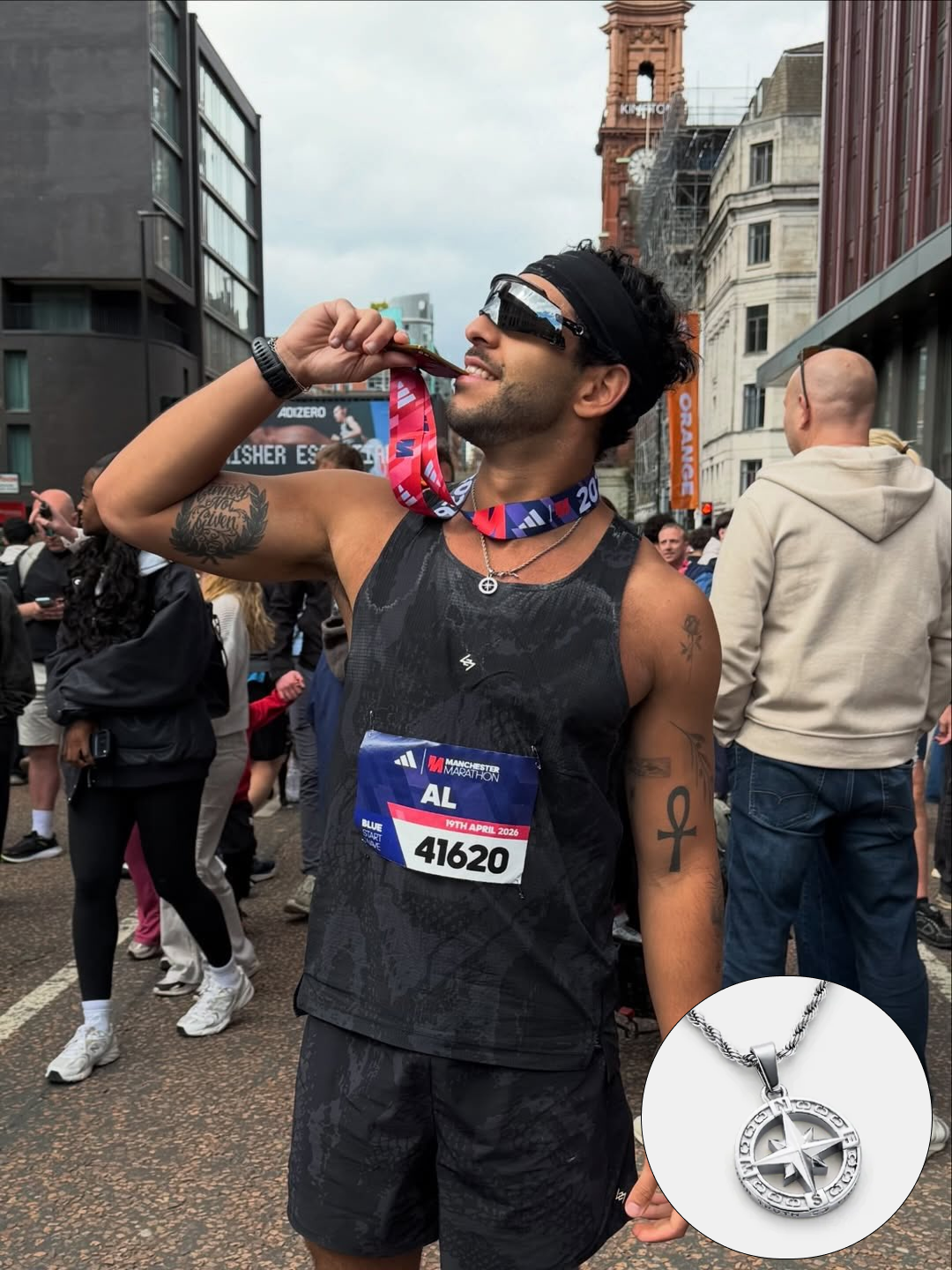 A male runner bites his Manchester Marathon medal while wearing sunglasses and a compass rose necklace.