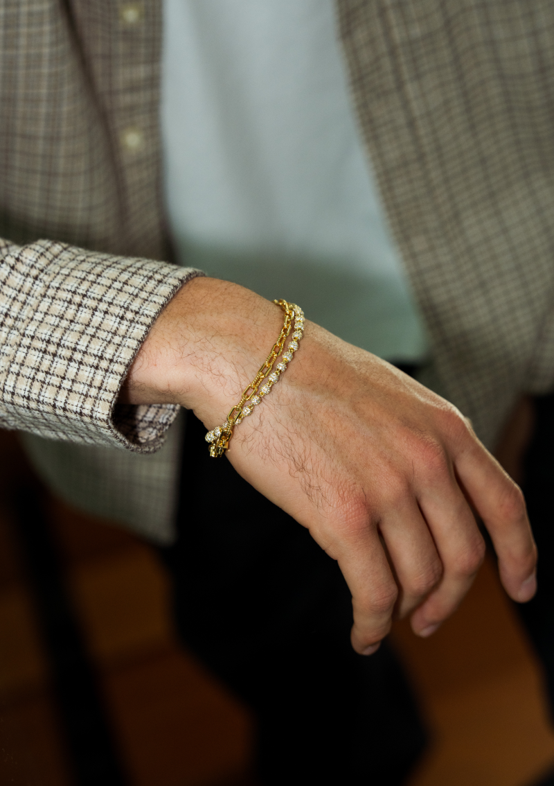 A close-up of a man's wrist, wearing a plaid blazer and two layered gold bracelets.
