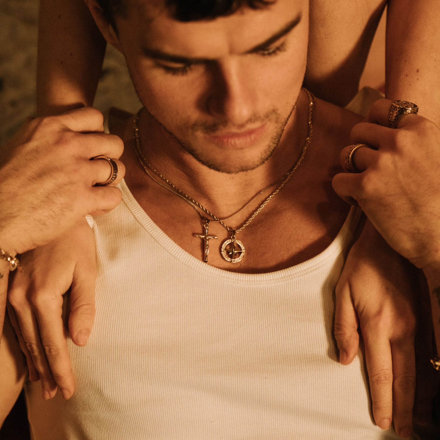 A man in a white tank top and gold jewelry is embraced from behind by another person.