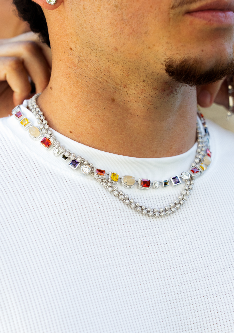A man wearing layered necklaces with colorful gemstones and diamonds over a white waffle-knit shirt.