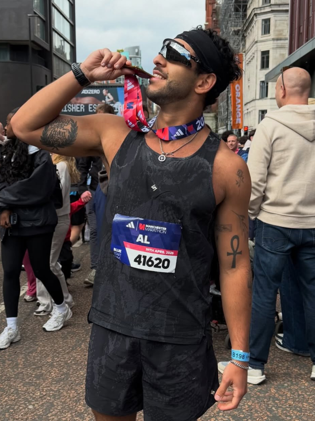 A male runner with tattoos smiles as he holds a Manchester Marathon medal to his mouth.