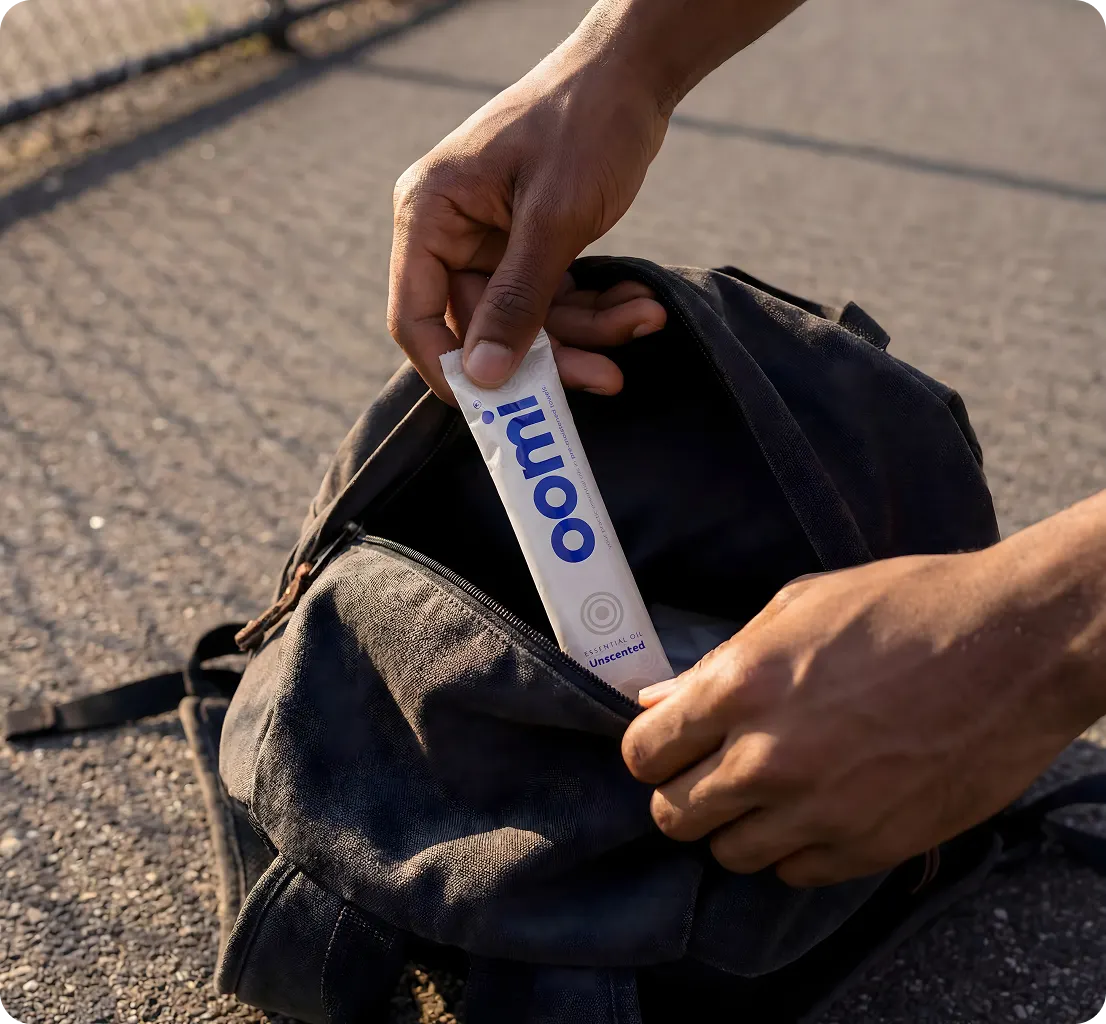 A person places a white Oomi packet into the front pocket of a black backpack.