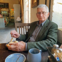 An older man with glasses sits at a table in a cafe, eating a meal.