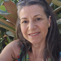 A headshot of a smiling woman with long, graying hair, with large-leafed plants in the background.