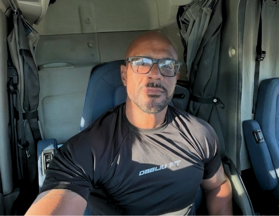 A bald man with glasses and a goatee sits in the driver's seat of a truck cab.