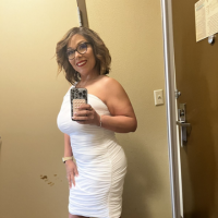 A woman with glasses and a white dress takes a selfie in a mirror.