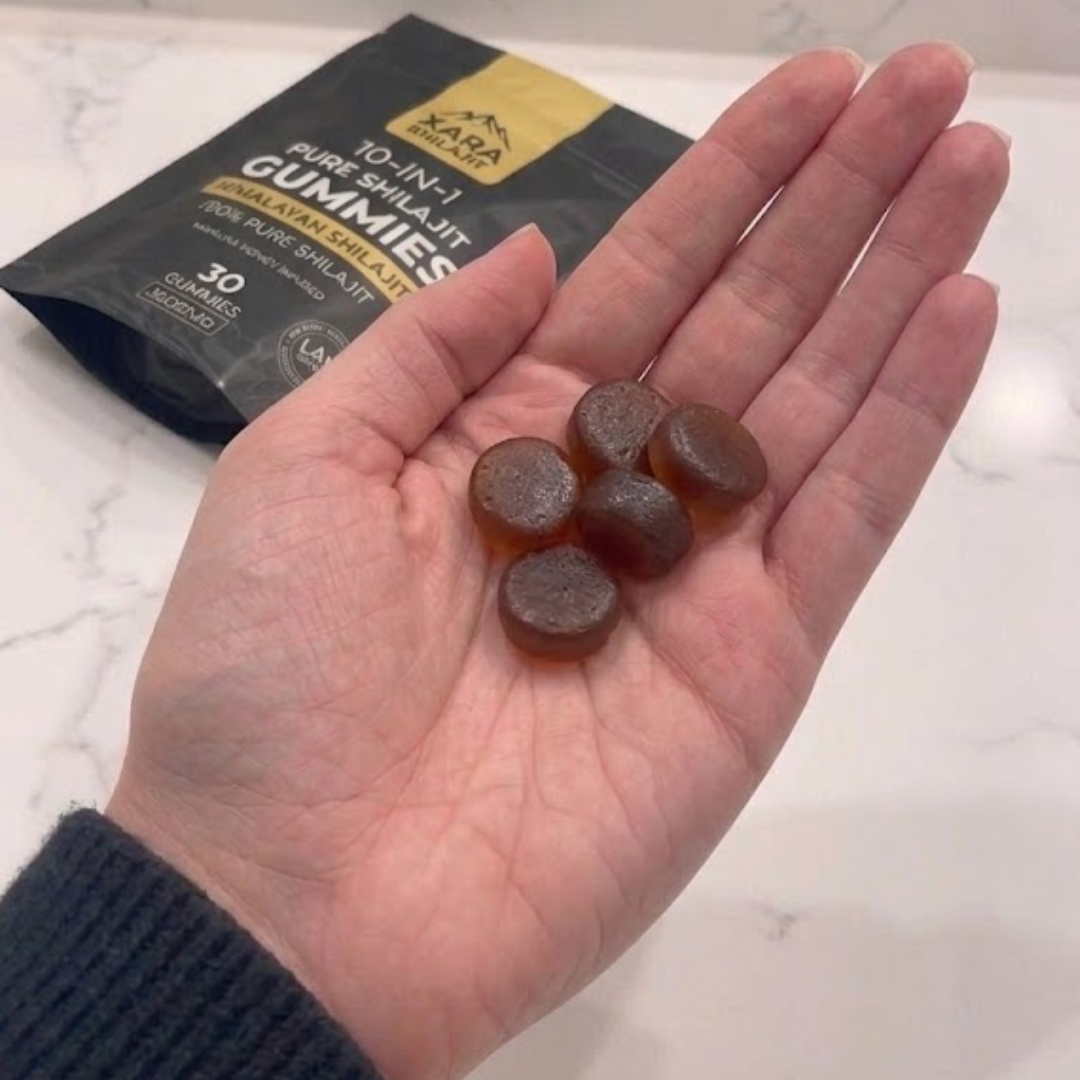 A hand holds five dark, round gummies with a pouch of Xara Shilajit Gummies in the background.