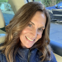 A smiling woman with long brown hair, wearing a blue jacket and sitting inside a car.