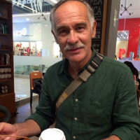 A man with a mustache and graying hair, wearing a green collared shirt and a bag strap.