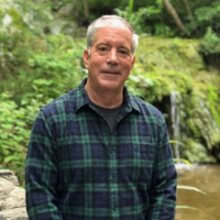 A man with short gray hair wearing a blue and green plaid shirt, standing in a garden.
