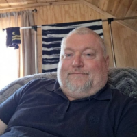 A middle-aged man with a gray beard and a blue polo shirt, smiling slightly in a wood-paneled room.